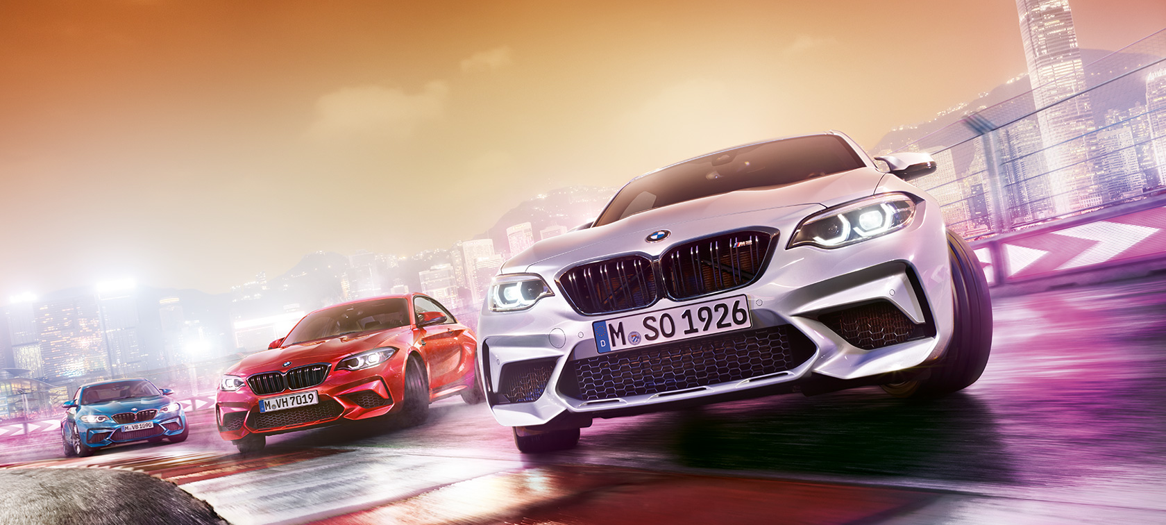 BMW M2 Competition in action Three BMW M2 Competition drifting on race track in front of nocturnal cityscape.
