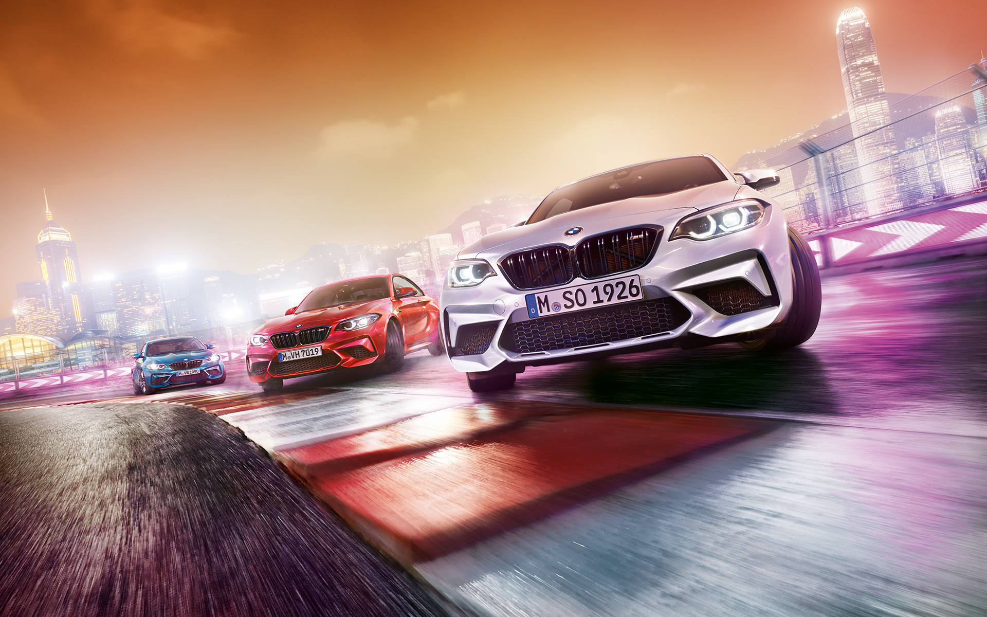 BMW M2 Competition in action Three BMW M2 Competition drifting on race track in front of nocturnal cityscape.