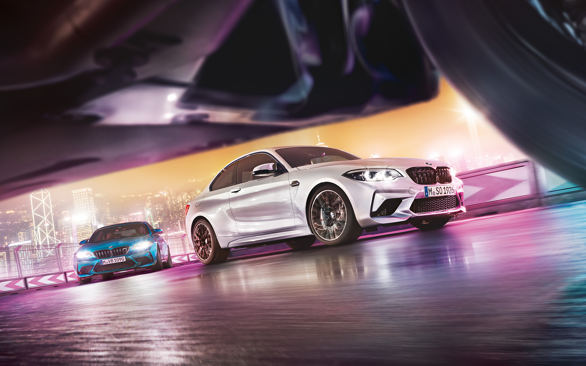 BMW M2 Competition on reflective asphalt Two BMW M2 Competition on race track in front of nocturnal cityscape in street-level view.