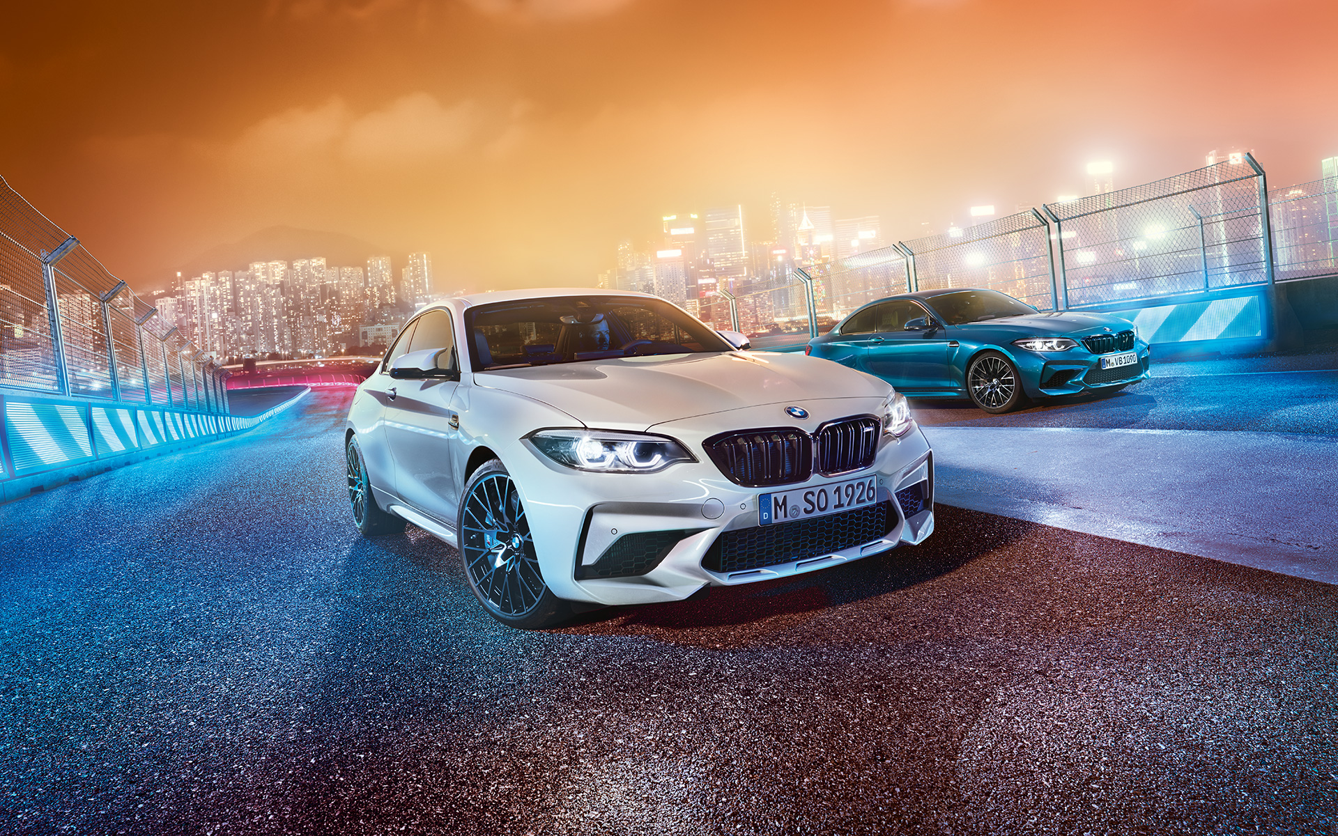 Two BMW M2 Competition on race track White and blue BMW M2 Competition in skewed front view standing in front of nocturnal cityscape.