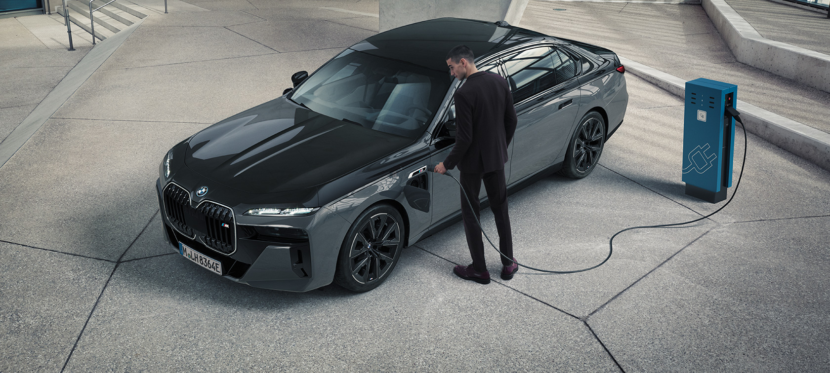 BMW M760e xDrive Sedan G70 M Performance 套件 BMW Individual 雙色 Black Sapphire 金屬漆面與 BMW Individual Dravite Grey 金屬漆面四分之三前視圖，一個男人正手持充電站旁的充電纜。